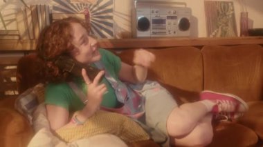 80 'ler tarzında iç tasarımla oturma odasında kanepede uzanmış, sabit telefondan konuşan, sakız çiğneyen kızıl saçlı beyaz kız.