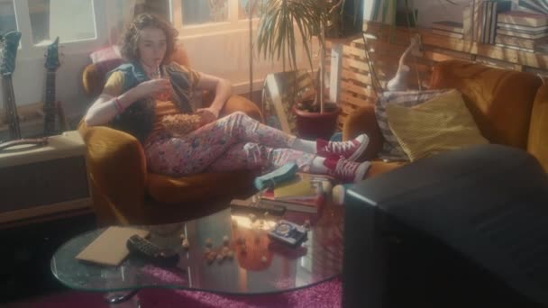 80年代スタイルのインテリアデザインと部屋のアームチェアに座っている若い白人の女の子の長いショット レトロテレビで映画を見ます ソーダを飲み ポップコーンを食べます — ストック動画