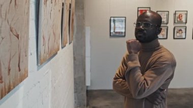 Modern sanat galerisinin duvarlarında resim serisi gözlemleyen genç ve zeki siyahi bir adamın belden yukarı bakış açısı.
