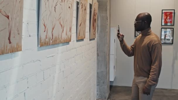 現代美術ギャラリーの壁に絵のシリーズのスマートフォンで写真を撮る若いインテリジェントブラックマンの中長期 — ストック動画