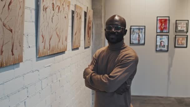 年轻聪明的黑人男子站在现代艺术馆墙上的一系列绘画的前景中 在镜头前微笑着看着 — 图库视频影像