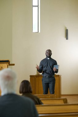 Katolik kilisesinde kürsüde dikilirken kilise cemaatiyle iletişim kuran rahip kılığındaki genç Afrikalı Amerikalı papaz.
