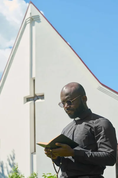 開いている聖書と木製のバラのビーズを持つ深刻なアフリカ系アメリカ人の男は教会の建物に対して福音から詩を読んで — ストック写真