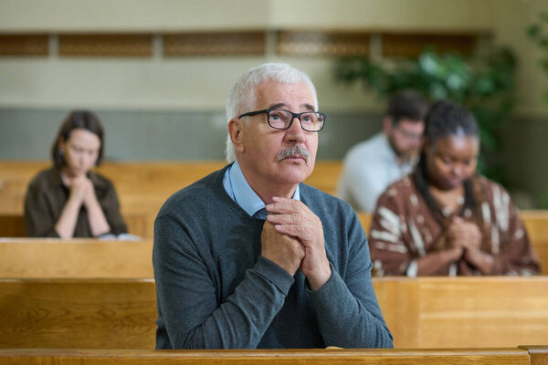Aged man in casualwear and eyeglasses keeping his hands put together during silent pray while looking at cross in church