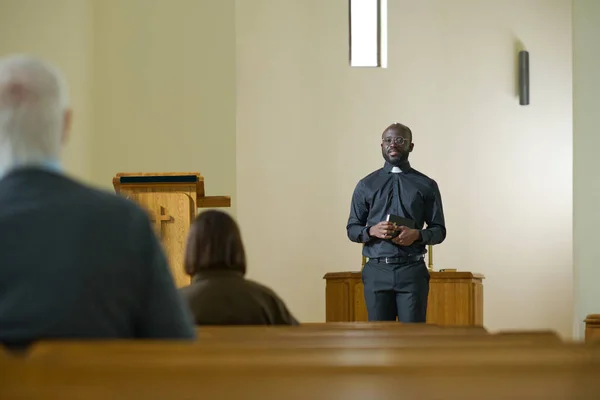 身穿牧师服装 头领的年轻黑人男子站在教区居民面前 在布道期间解释 中的诗句 — 图库照片