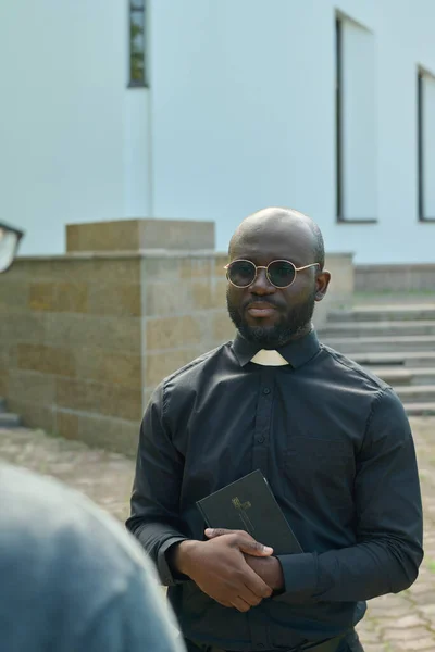教会の庭に白い建物に対して立っている間聖職襟のコンサルティング教区と黒のシャツの若いアフリカ系アメリカ人牧師 — ストック写真