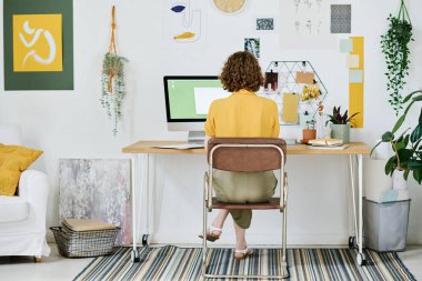 Serbest giyinen genç bir kadın bilgisayar monitörünün önünde iş yerinde oturup evde yeni web siteleri yaratıyor.