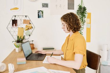 Gündelik kıyafet giymiş genç ciddi bir iş kadını dizüstü bilgisayarın önünde oturup finansal belgelere not alıyor ve verileri analiz ediyor.