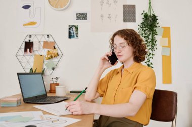 İş yerinde bilgisayar ve mali belgelerle otururken akıllı telefonlu genç bir kadın iş organize ediyor ve müşterileri arıyor.