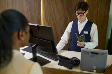 Üniformalı ve gözlüklü genç bir Asyalı otel odasında önünde duran misafirin pasaportunda kişisel bilgilerini kontrol ediyor.