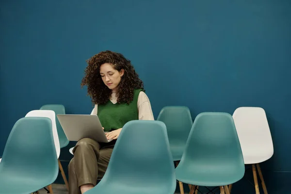 年轻的黑发女商人 留着波浪般的头发 坐在空荡荡的课室和网络中的一张椅子上 在笔记本电脑键盘上打字 — 图库照片
