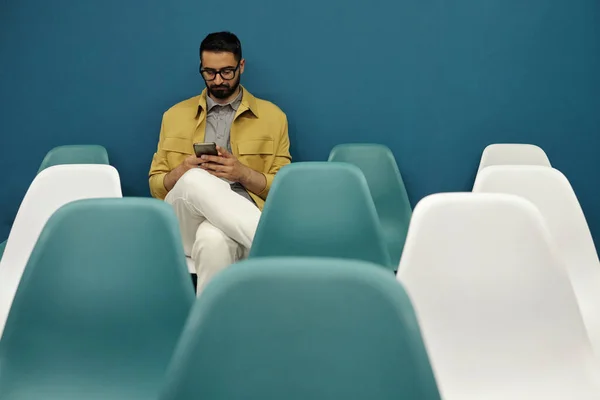年轻的胡子商人穿着便服 戴着眼镜 坐在讲台上的一张椅子上 一边用智能手机发短信 — 图库照片
