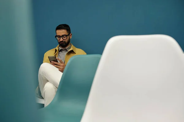 认真的商人 坐在白椅和蓝椅子之间的讲台上 拿着智能手机浏览在线数据或照片 — 图库照片