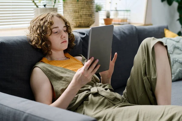 若いですリラックスした白人女性でカジュアルウェアで自宅でソファに休んでいる間デジタルタブレットの画面上のオンラインデータを見て — ストック写真