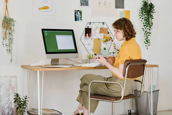 Young serious businesswoman scrolling in mobile phone while sitting in front of computer monitor and organizing work at home