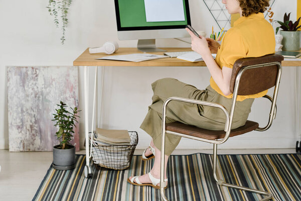 Cropped shot of young freelancer in casualwear sitting on chair in front of computer monitor by workplace and scrolling in mobile phone