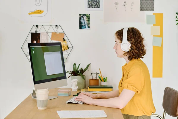 年轻的现代自由职业者坐在带有小空白窗口的监视器前 在计算机键盘上打字的侧视图 — 图库照片