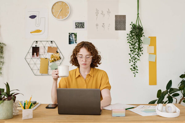 Young serious freelancer with cup of tea sitting in front of laptop by workplace and searching for online information for project