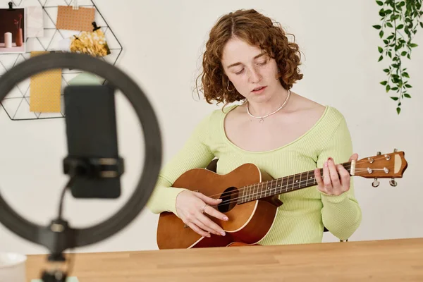 Young female student or teacher of course of guitar playing sitting by desk in front of smartphone camera during online lesson