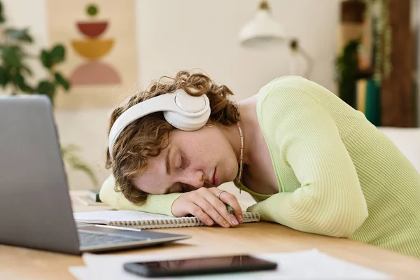 年轻疲惫的女学生头戴耳机 穿着便服 一边在笔记本电脑前的桌子旁打盹 一边把脑袋贴在复印机上 — 图库照片