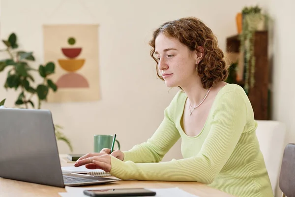 年轻自信的女商人坐在笔记本电脑前的桌子旁 一边做笔记 一边浏览在线信息 — 图库照片