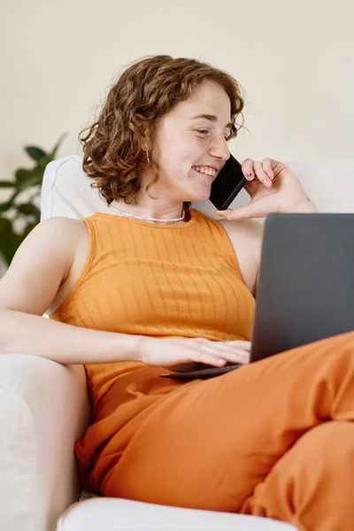 坐在舒适的扶手椅上 双膝跪在笔记本电脑上 一边用手机说话的快乐的年轻女商人 — 图库照片