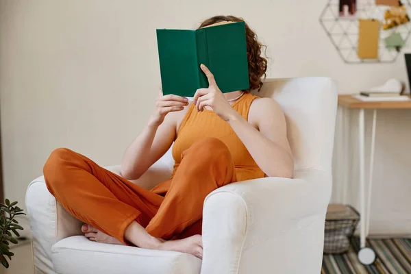 若いです女性でオレンジ色のカジュアル読書本でダークグリーンカバーしながら座っている間に白い革快適なアームチェアでリビングルーム — ストック写真