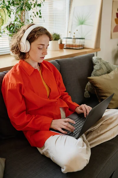 年轻而认真的女商人或戴耳机和便服的女学生 双腿交叉 关系网 坐在沙发上 手持笔记本电脑 — 图库照片