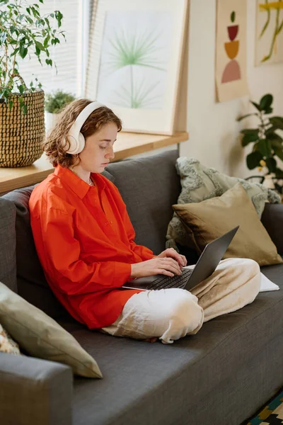 若いです女性学生で赤いシャツと白いパンツタイプのラップトップキーボード上で座っている間ソフト快適なソファでリビングルーム — ストック写真