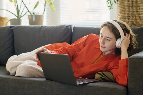 若いです女の子でヘッドフォンの上に横たわるソフト快適なソファでリビングルームとラップトップ画面を見ながらオンライントレーニング — ストック写真
