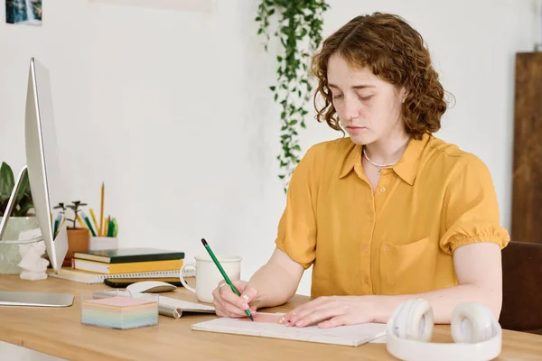 年轻富有创造力的女自由撰稿人 坐在电脑监控器前的桌子旁边 手握尺子和铅笔 在抄本上画草图 — 图库照片