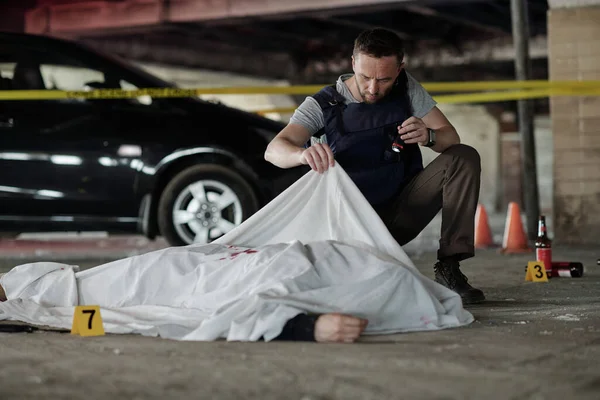 駐車場で犯罪現場に横たわっている死体を検査しながら小さなランタンを保持防弾チョッキの若い男性犯罪学の専門家 — ストック写真