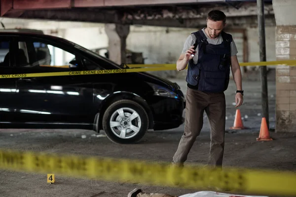 駐車場のアスファルトに横たわっている死体によって立っている間 若い男性犯罪現場の捜査官は殺人の証拠を探しています — ストック写真