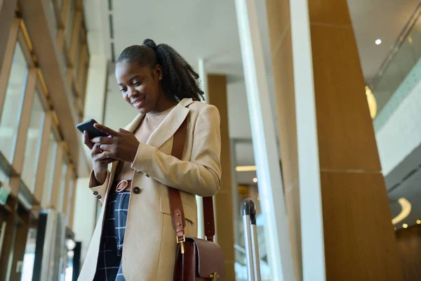 年轻微笑的黑人女人站在酒店走廊或休息室时 用智能手机发短信或与朋友通过视频聊天 — 图库照片