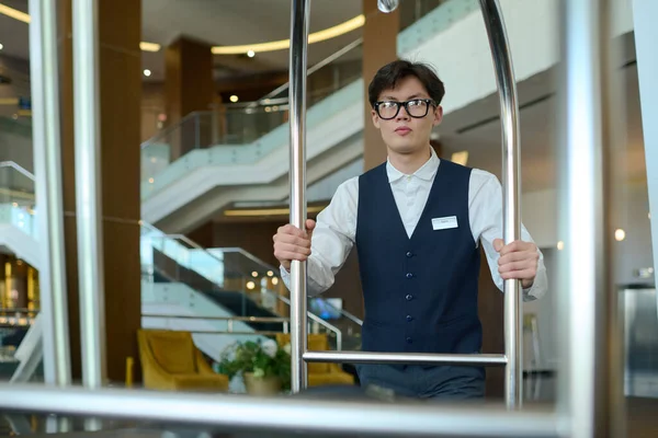 アジアの民族の若い深刻な男が正式に進む廊下に沿って移動しながら ホテルのクライアントのスーツケースとカートを押します — ストック写真