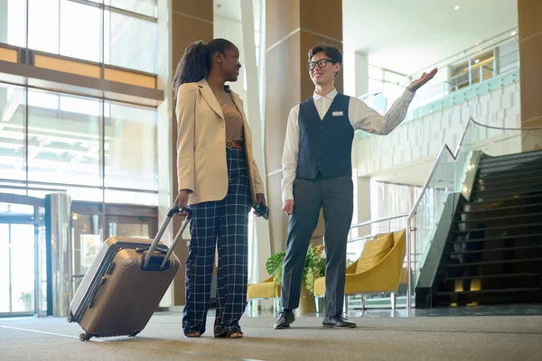 現代的なホテルの若い笑顔のマネージャースーツケースを持つ女性旅行者がラウンジに入り 受付カウンターに向かって移動します — ストック写真