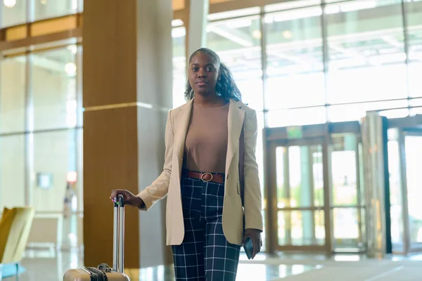 年轻而严肃的非洲裔美国女人穿着时髦的便服 站在宽敞奢华的宾馆里 手握手提箱 — 图库照片