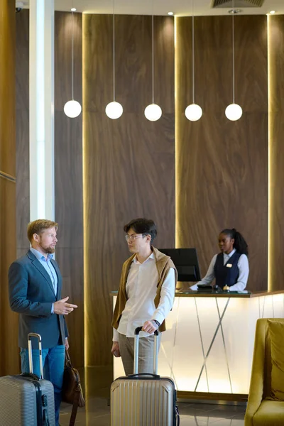 ホテルのラウンジで受付カウンターに立ち 旅の仕事のポイントを話し合う2人の若いエレガントなビジネスマン — ストック写真
