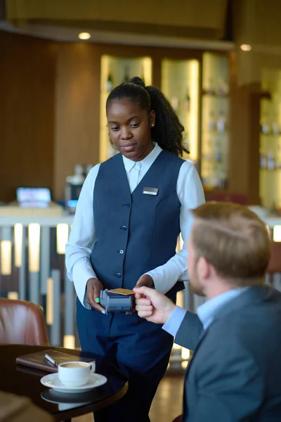 若いアフリカ系アメリカ人女性ホテルの労働者の支払い端末のビジネスマンの前でコーヒーのクレジットカードで支払う立っている — ストック写真
