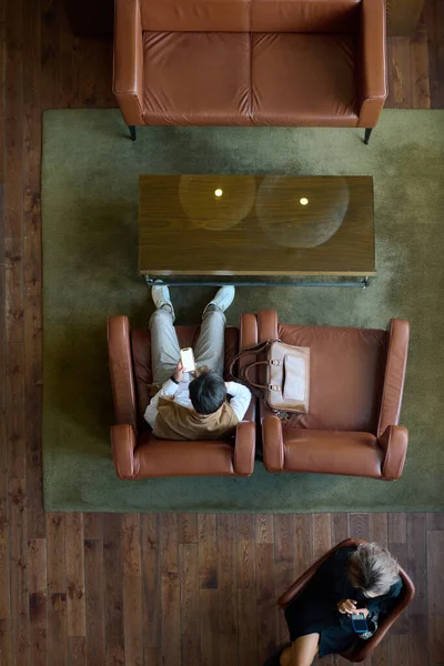 图为两个年轻的商人坐在现代宾馆的沙发上 一边用手机发短信一边聊天的情景 — 图库照片