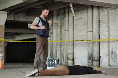 Kurşun geçirmez yelek giyen genç dedektif ya da polis olay yerini incelerken cesedin yanında duruyor.
