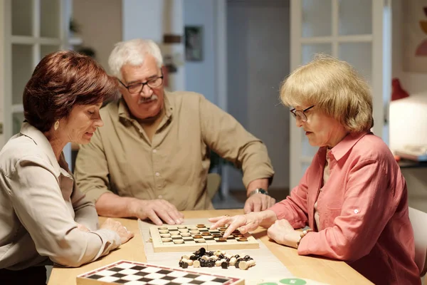 チェスの試合のためにリビングルームでテーブルによって集められた3人のシニアの友人 何木製の図を移動することを考えている高齢女性 — ストック写真