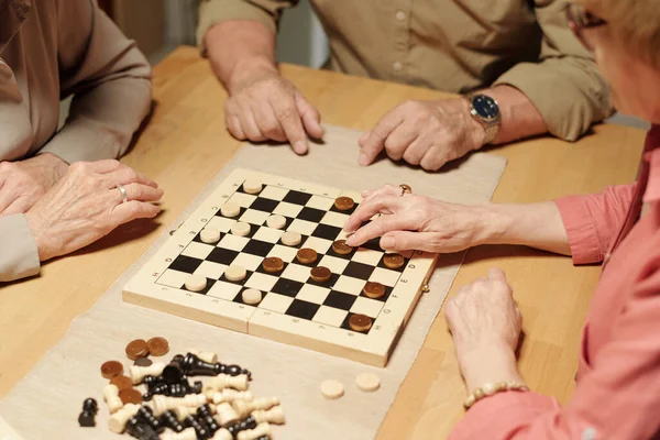Hands of three senior friendly people gathered by table with checkers, chess figures and chessboard to play leisure games