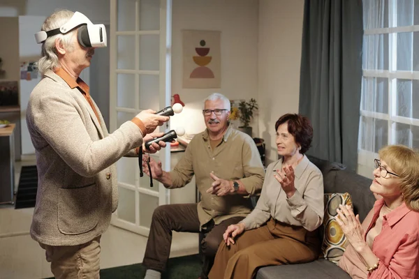 stock image Senior man in vr headset controlling picture on screen of tv set with two gamepads while standing in front of his friends sitting on sofa