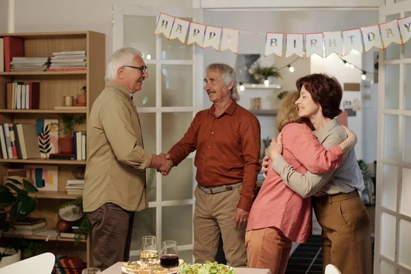 两个年长的女人互相拥抱 而她们的丈夫则在客厅里和准备生日派对的餐桌握手 — 图库照片