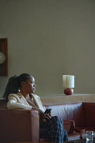 年轻而安静的非洲裔美国女商人 穿着时髦的便衣 坐在宾馆的皮革沙发上 用智能手机 — 图库照片