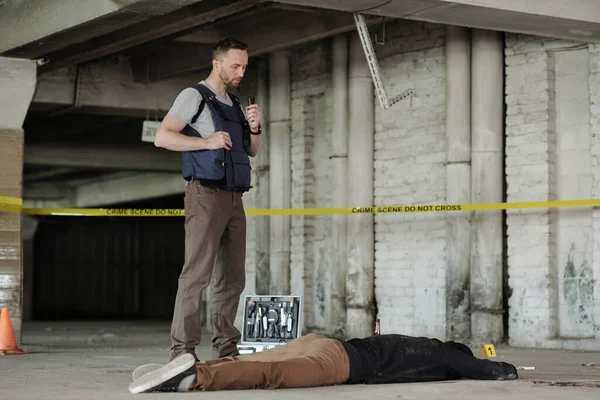 穿着防弹背心站在尸体旁边检查犯罪现场的年轻男性侦探调查员或警察 — 图库照片
