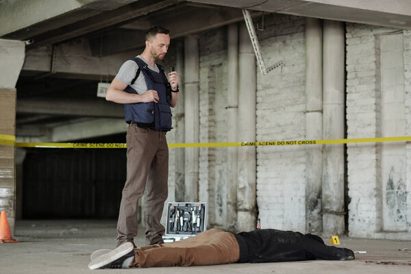 Young male detective investigator or policeman in bulletproof vest standing by dead body while inspecting crime scene