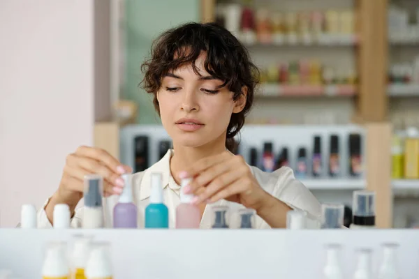 Young assistant of cosmetic shop putting new self care products in small plastic bottles on display while working in cosmetic shop