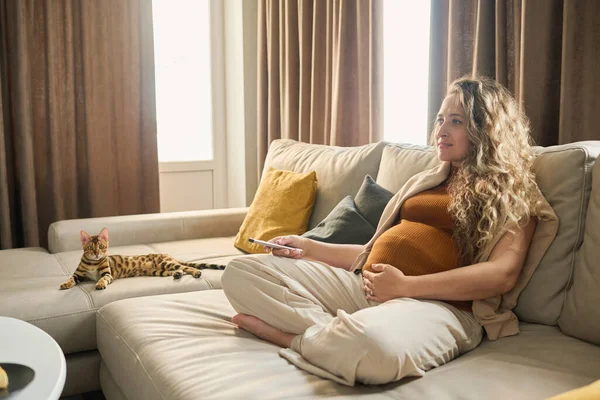 年轻而安静的孕妇穿着便服 穿着遥控器上的按键 与纯洁的宠物在沙发上悠闲自在地呆在家里 — 图库照片